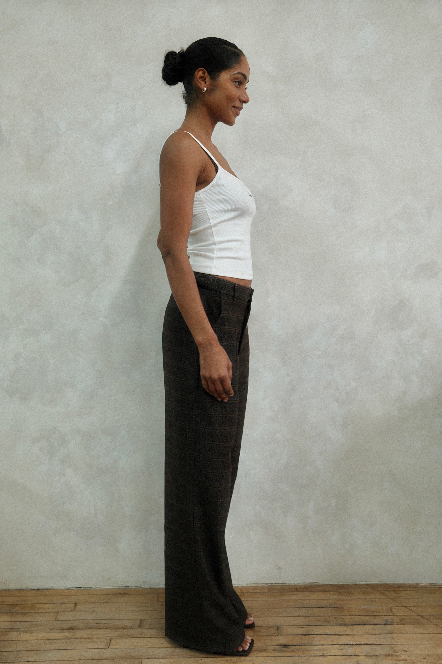 Side profile of model wearing brown windowpane plaid Carter Pants with white LÉLA tank top and gold hoop earrings against textured white wall
