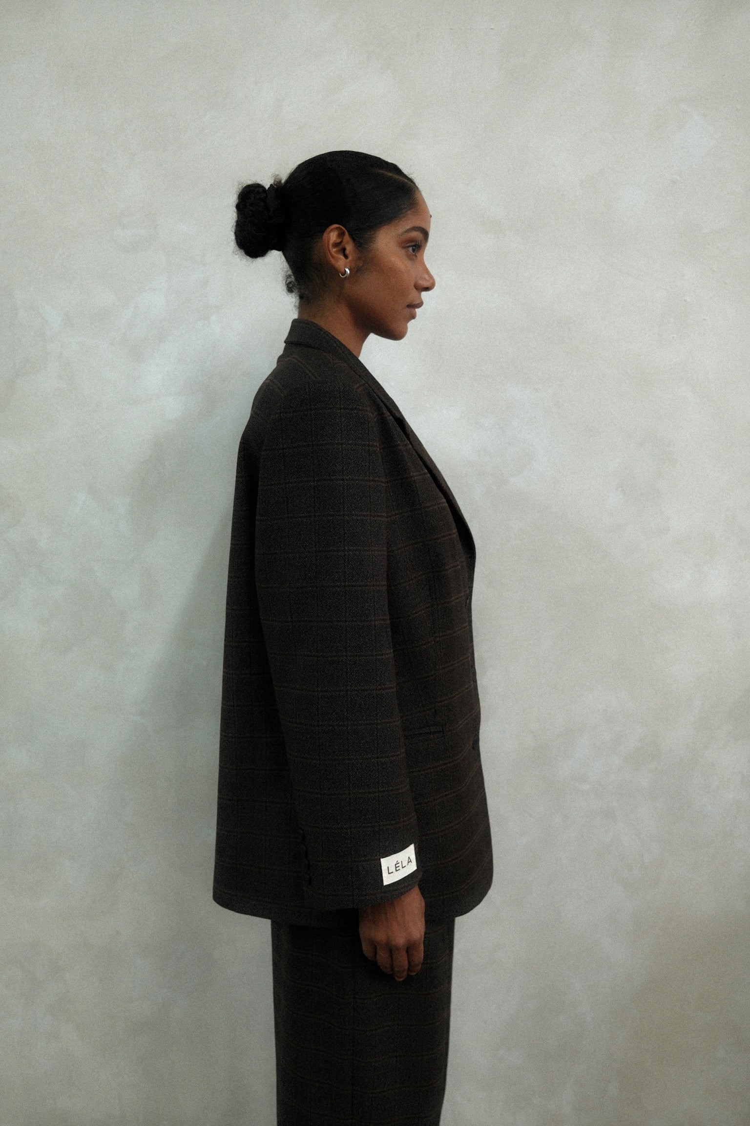 Side view of model wearing brown windowpane plaid Archer Blazer with white LÉLA branded label visible on cuff, styled with matching pants