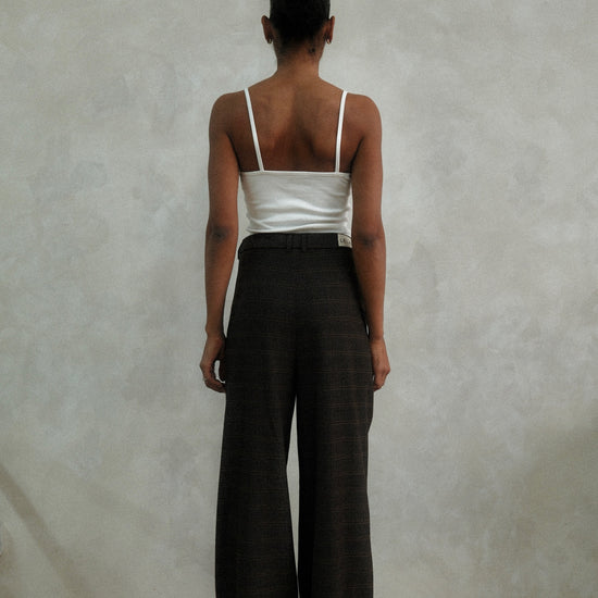 Back view of model wearing brown windowpane plaid Carter Pants with white ribbed tank top, showing wide-leg silhouette against textured white wall