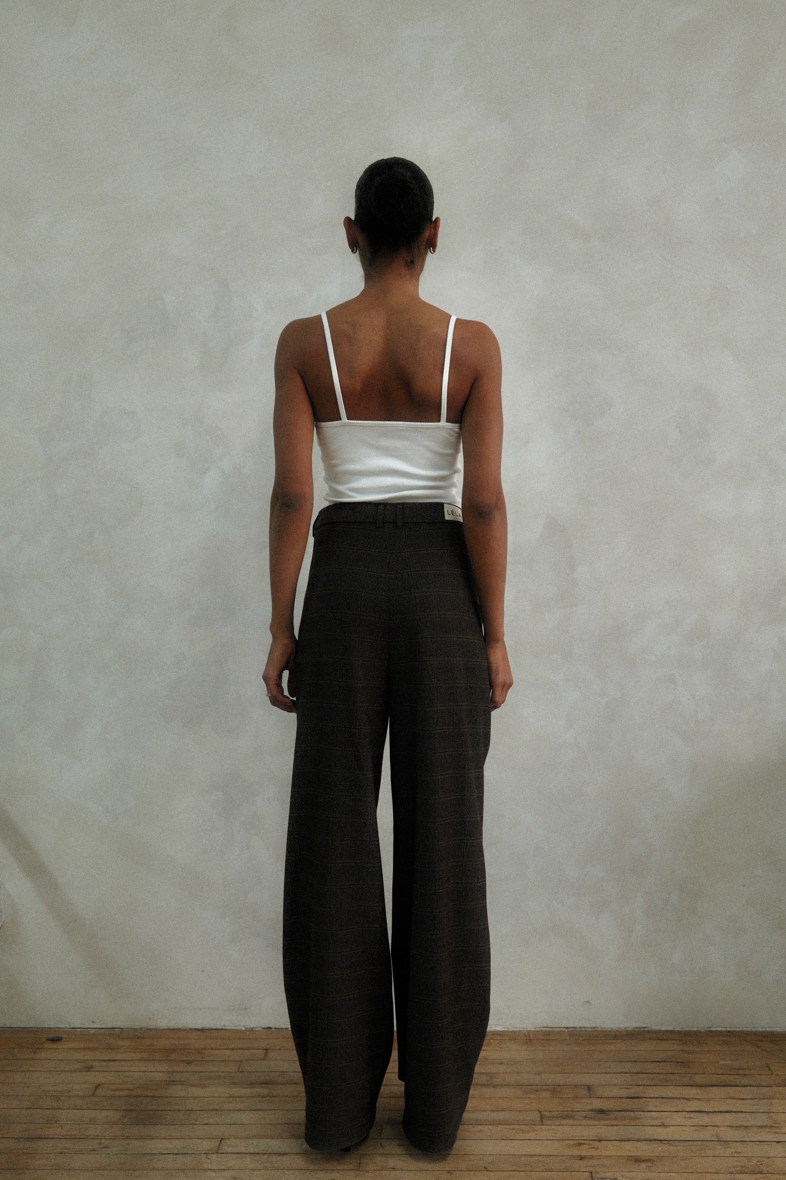 Back view of model wearing brown windowpane plaid Carter Pants with white ribbed tank top, showing wide-leg silhouette against textured white wall