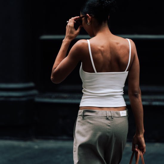 Back view of a woman walking away wearing the white ribbed LÉLA tank top and beige LÉLA Archer trousers with a LÉLA label on the rear waistband, holding a brown leather handbag