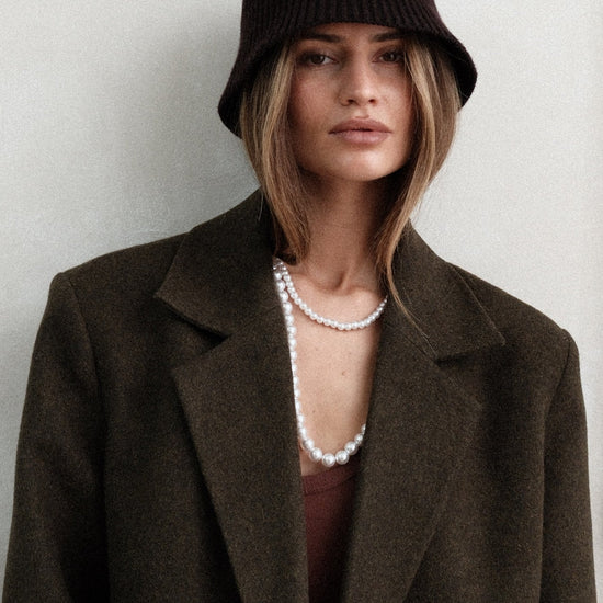 Model wearing dark green Beaumont Coat with brown bucket hat and pearl necklace, styled with brown tank top, gloves and dark washed jeans against white background