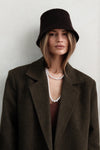 Model wearing dark green Beaumont Coat with brown bucket hat and pearl necklace, styled with brown tank top, gloves and dark washed jeans against white background