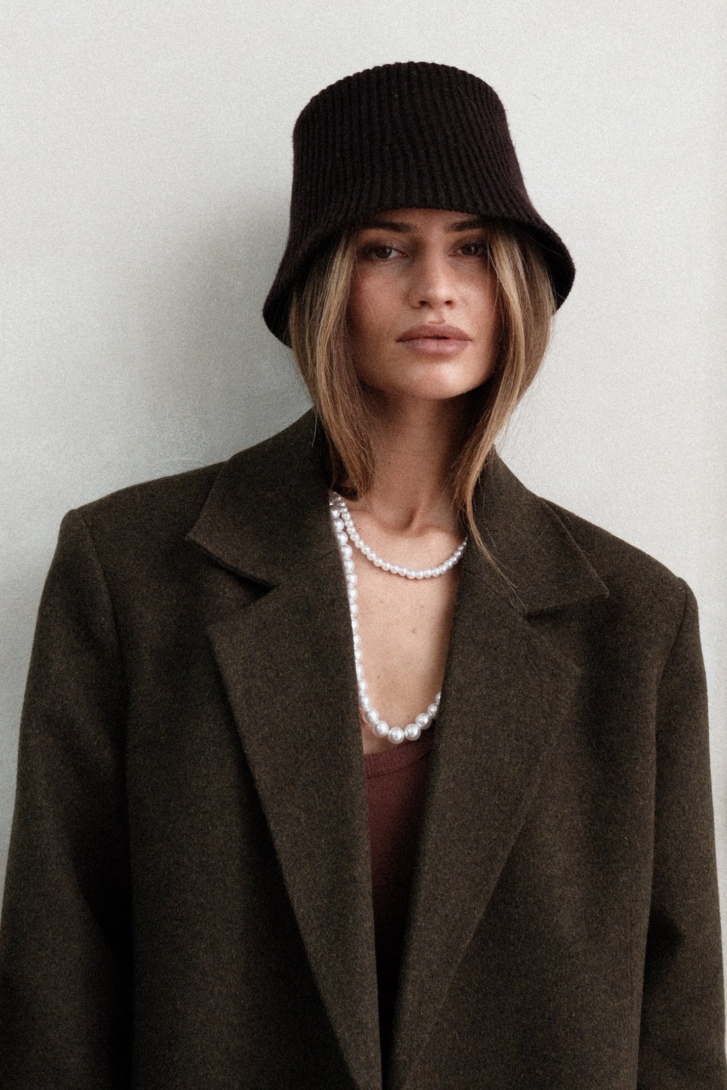 Model wearing dark green Beaumont Coat with brown bucket hat and pearl necklace, styled with brown tank top, gloves and dark washed jeans against white background