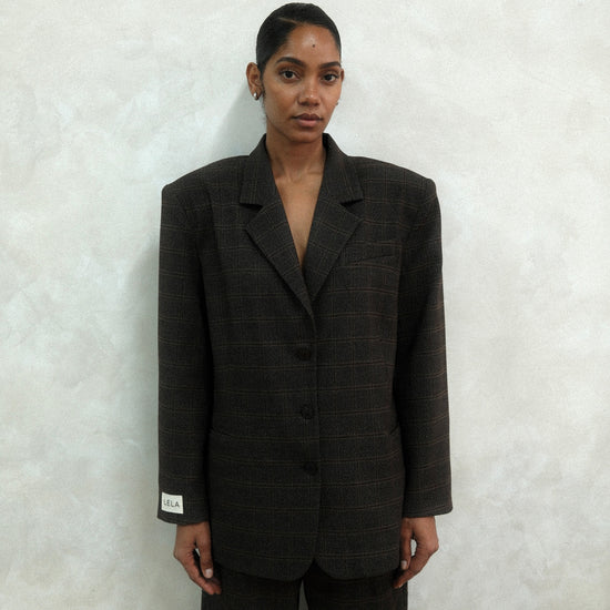 Front view of model wearing brown windowpane plaid Archer Blazer with white LÉLA label on cuff, styled with matching pants against textured white wall