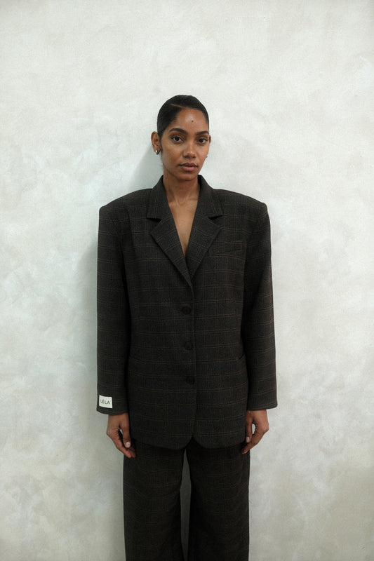Front view of model wearing brown windowpane plaid Archer Blazer with white LÉLA label on cuff, styled with matching pants against textured white wall