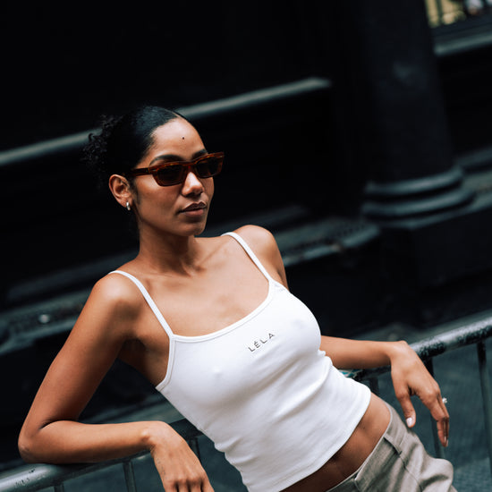 Woman leaning back against a metal railing in an urban setting, wearing rectangular sunglasses, the white LÉLA tank top with brown embroidery, and beige LÉLA Carter trousers