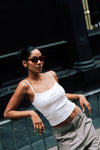 Woman leaning back against a metal railing in an urban setting, wearing rectangular sunglasses, the white LÉLA tank top with brown embroidery, and beige LÉLA Carter trousers
