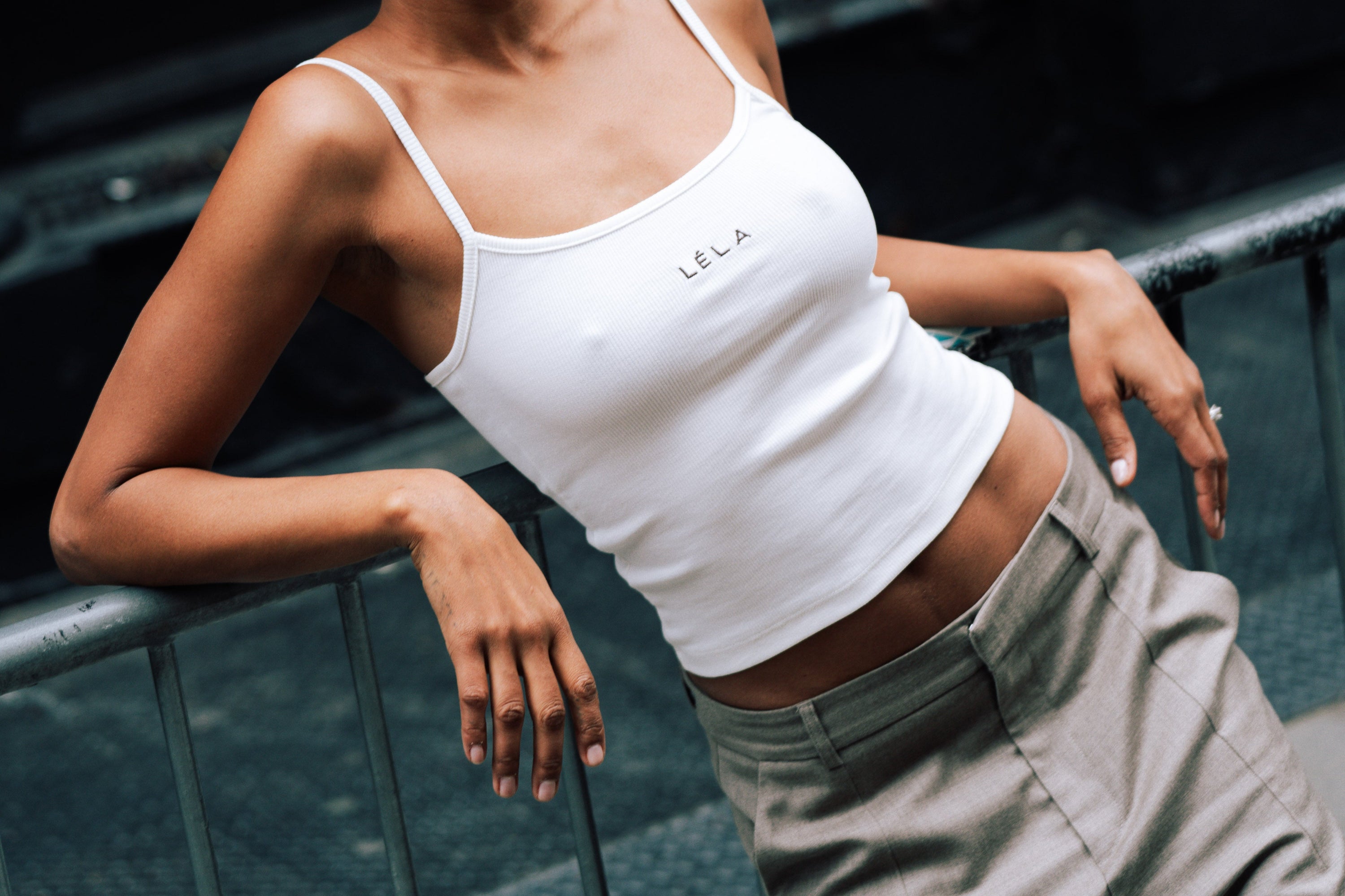 Woman leaning back against a metal railing in an urban setting, wearing rectangular sunglasses, the white LÉLA tank top with brown embroidery, and beige LÉLA Carter trousers