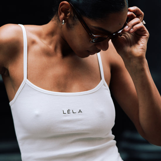 Close-up of a woman wearing the white ribbed LÉLA tank top with brown LÉLA embroidered on the chest, adjusting her tortoiseshell sunglasses