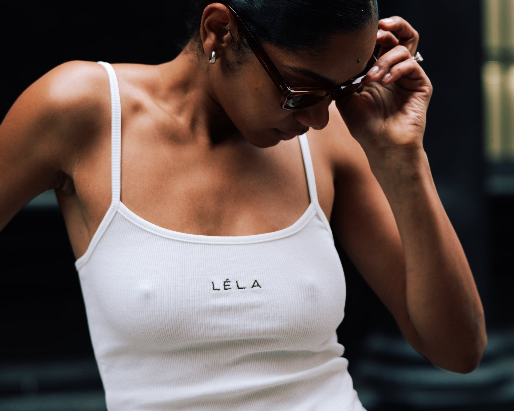 Close-up of a woman wearing the white ribbed LÉLA tank top with brown LÉLA embroidered on the chest, adjusting her tortoiseshell sunglasses