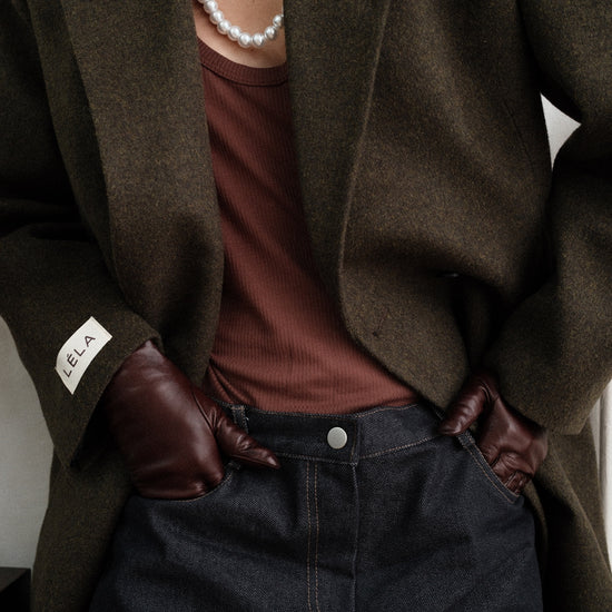 Close-up detail of dark green Beaumont Coat showing LÉLA branded label on cuff, styled with pearl necklace, brown tank top and gloves, and dark washed denim