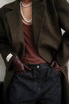 Close-up detail of dark green Beaumont Coat showing LÉLA branded label on cuff, styled with pearl necklace, brown tank top and gloves, and dark washed denim