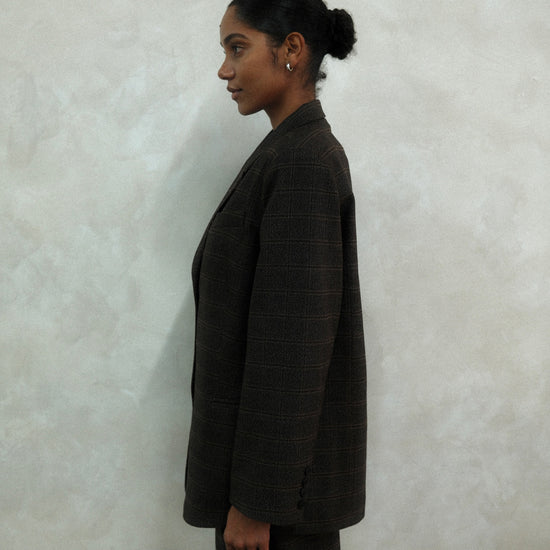 Side profile of model wearing brown windowpane plaid Archer Blazer with matching pants against textured white wall, showing oversized tailored silhouette