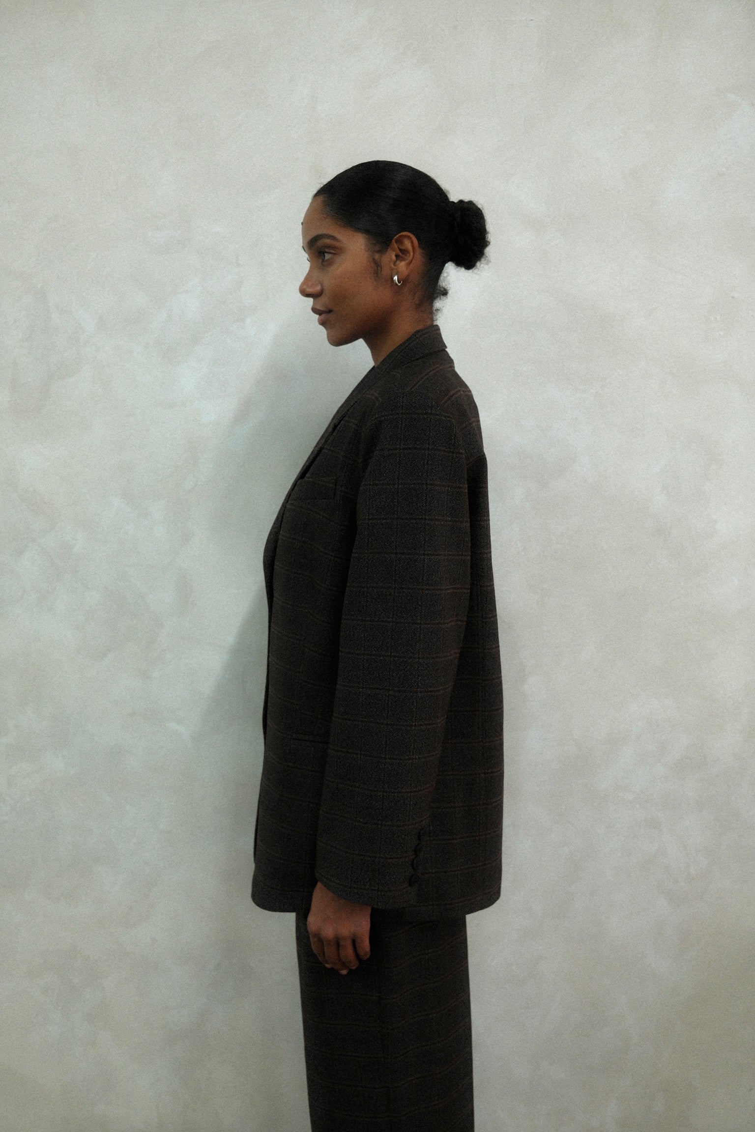 Side profile of model wearing brown windowpane plaid Archer Blazer with matching pants against textured white wall, showing oversized tailored silhouette