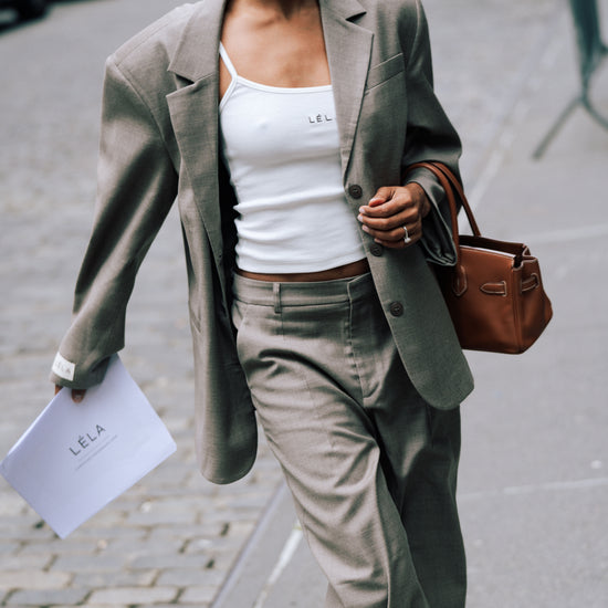 Model wearing beige Carter Blazer with matching pants, white LÉLA tank top, brown sunglasses and brown leather handbag on city street, carrying a LÉLA-labeled sheet