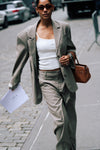 Model wearing beige Carter Blazer with matching pants, white LÉLA tank top, brown sunglasses and brown leather handbag on city street, carrying a LÉLA-labeled sheet