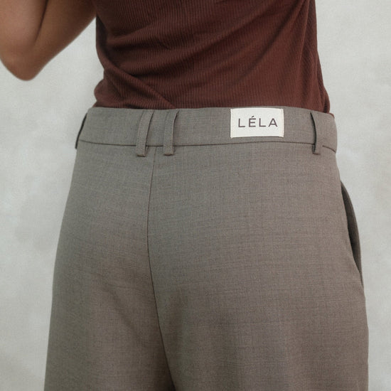 Back detail view of beige Carter Pants showing LÉLA branded label on waistband, styled with brown tank top against white background