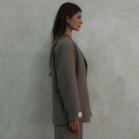 Side profile of model wearing beige Carter Blazer with matching pants and LÉLA label visible on cuff against textured white background