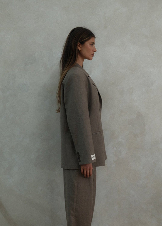Side profile of model wearing beige Carter Blazer with matching pants and LÉLA label visible on cuff against textured white background