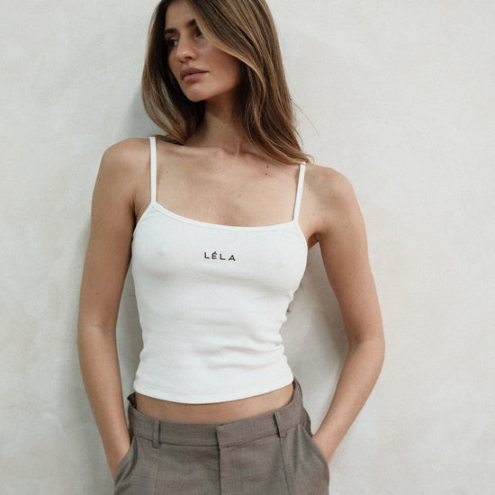 Woman standing against a neutral wall looking to the side, wearing the white LÉLA tank top and beige LÉLA Carter trousers with her hands in her pockets