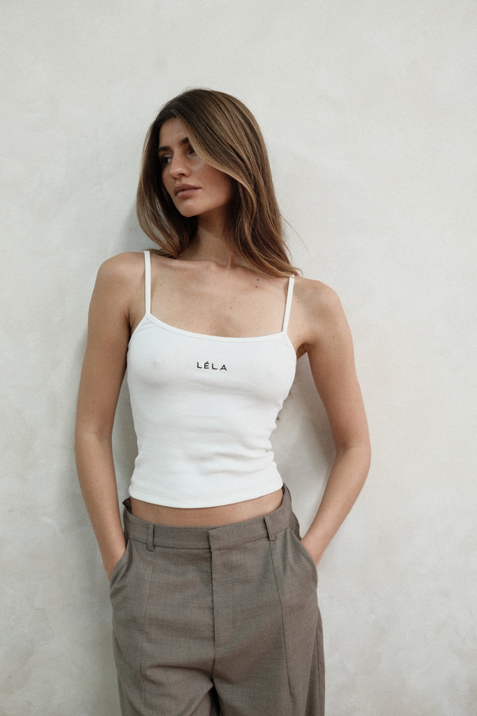 Woman standing against a neutral wall looking to the side, wearing the white LÉLA tank top and beige LÉLA Carter trousers with her hands in her pockets