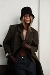 Model wearing dark green Beaumont Coat with brown bucket hat and pearl necklace, styled with brown tank top, gloves and dark washed jeans against white background