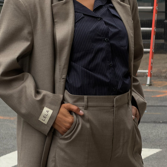 Close-up detail of beige Carter Blazer showing LÉLA branded label on cuff, styled with matching pants and blue pinstripe shirt in urban setting
