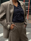 Close-up detail of beige Carter Blazer showing LÉLA branded label on cuff, styled with matching pants and blue pinstripe shirt in urban setting