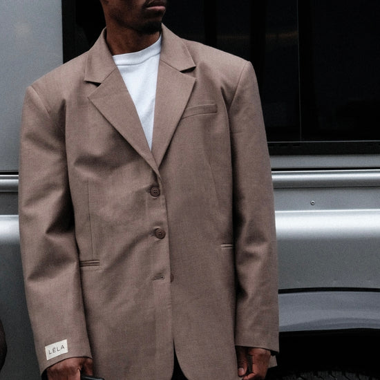 Close-up of male model wearing beige Carter Blazer with black sunglasses and white LÉLA label on cuff, standing by silver van