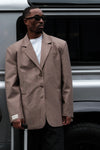 Close-up of male model wearing beige Carter Blazer with black sunglasses and white LÉLA label on cuff, standing by silver van