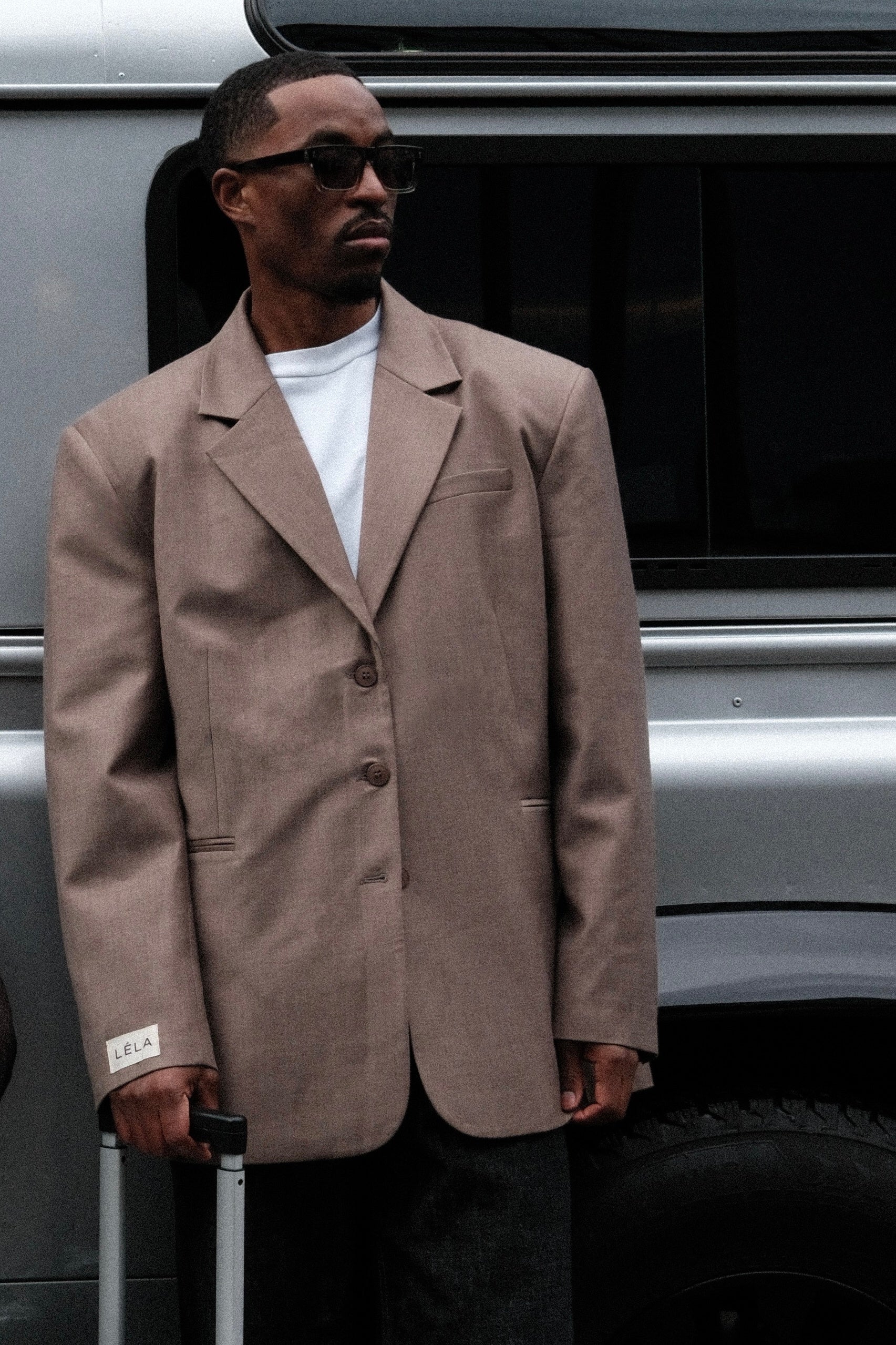 Close-up of male model wearing beige Carter Blazer with black sunglasses and white LÉLA label on cuff, standing by silver van