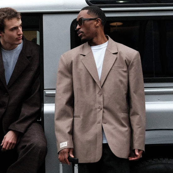 Male Model wearing beige Carter Blazer with black sunglasses, white t-shirt and silver luggage, standing by silver van with another person on his right