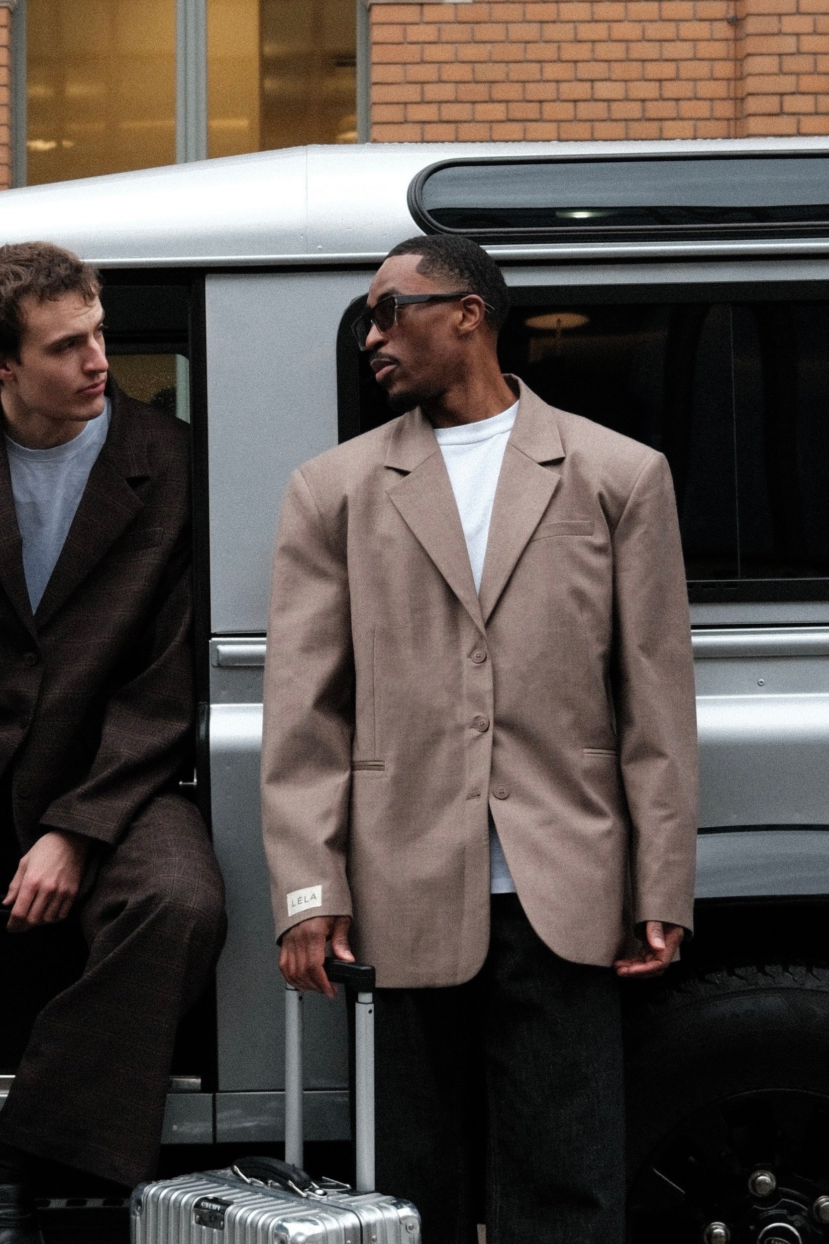 Male Model wearing beige Carter Blazer with black sunglasses, white t-shirt and silver luggage, standing by silver van with another person on his right