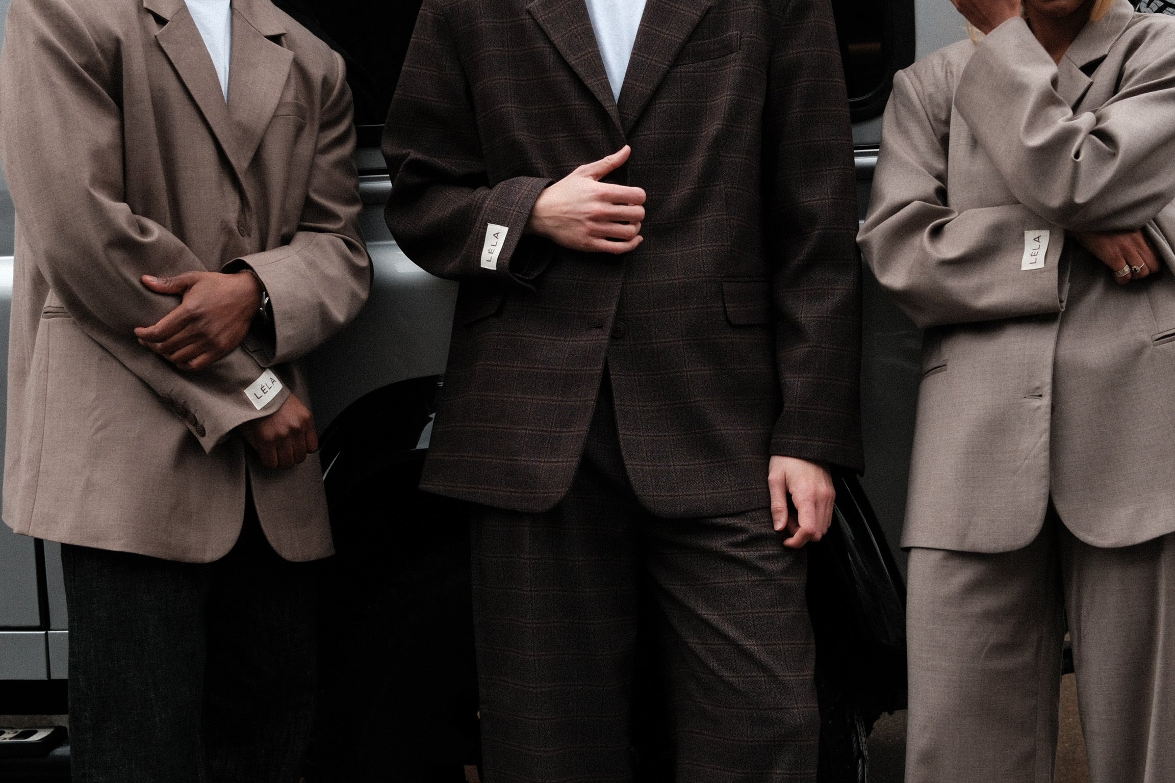 Three models wearing LÉLA Studios tailored suits in beige and dark brown windowpane check patterns with branded cuff labels