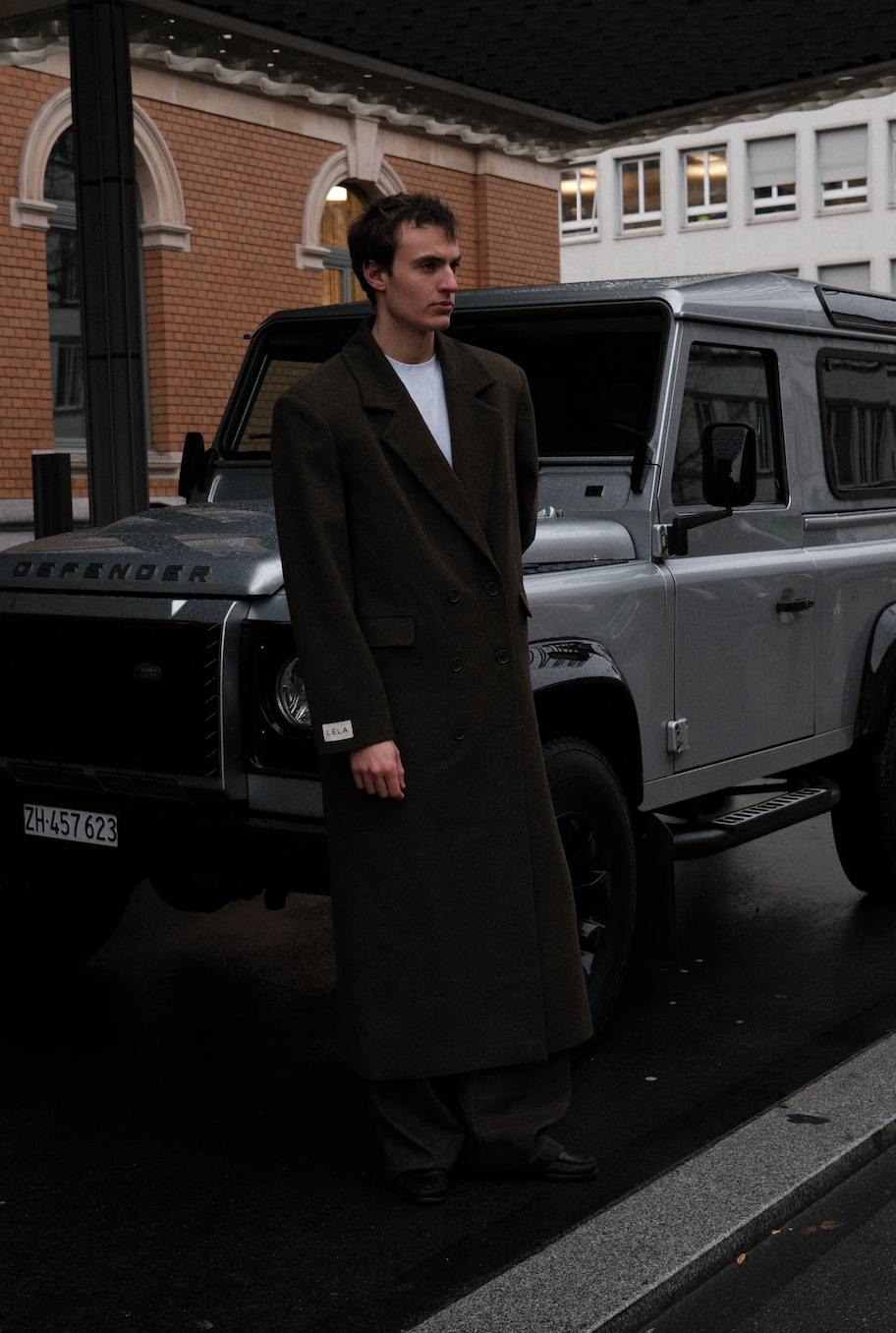Full-length view of model wearing dark green Beaumont Coat with white t-shirt, leaning against silver Land Rover Defender