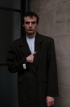Male model wearing dark green double-breasted Beaumont Coat with LÉLA label, styled with white t-shirt in urban concrete setting