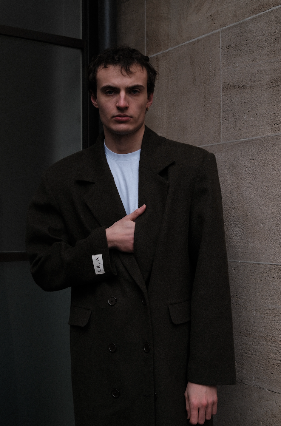 Male model wearing dark green double-breasted Beaumont Coat with LÉLA label, styled with white t-shirt in urban concrete setting