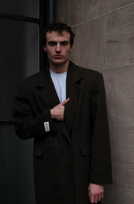 Male model wearing dark green double-breasted Beaumont Coat with LÉLA label, styled with white t-shirt in urban concrete setting