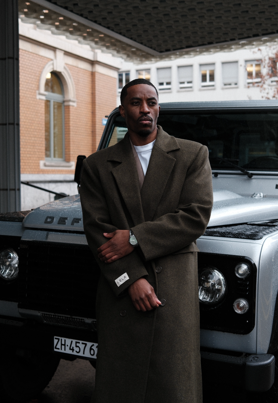 Full-length view of model wearing dark green Beaumont Coat white t-shirt, leaning against silver Land Rover Defender