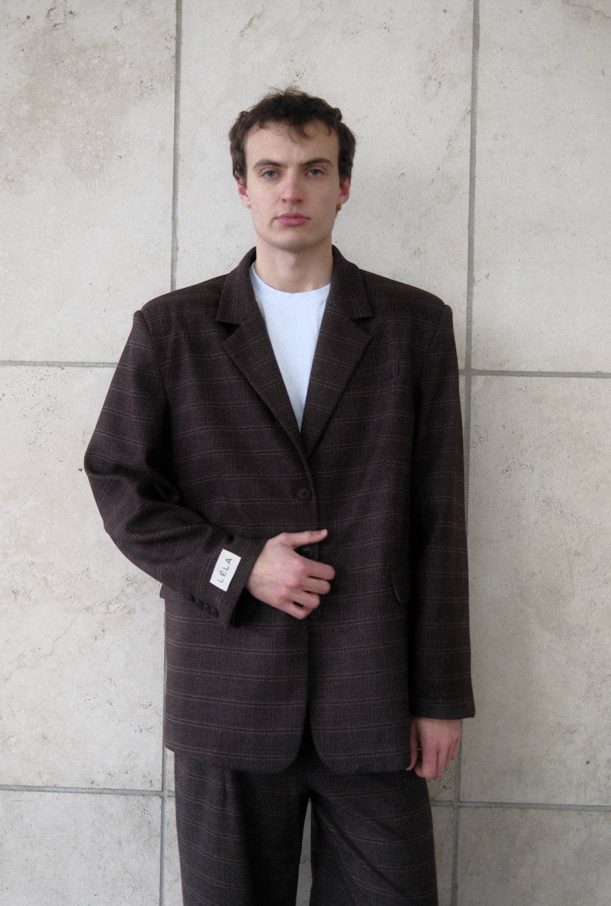 Male model wearing brown windowpane plaid Archer Blazer with white LÉLA label on cuff and white t-shirt, styled with matching pants against concrete wall
