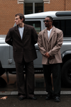 Left male model wearing brown plaid Carter Blazers with matching pants and white t-shirt, standing in front of silver Land Rover Defender against brick building with other male model to his left