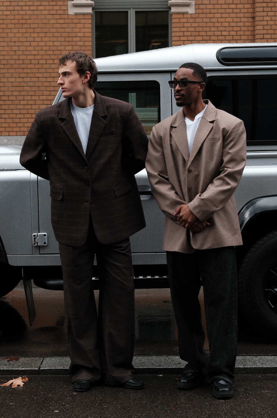 Left male model wearing brown plaid Carter Blazers with matching pants and white t-shirt, standing in front of silver Land Rover Defender against brick building with other male model to his left