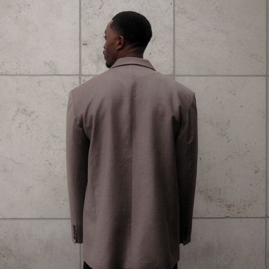 Back view of beige Carter Blazer showing tailored construction and length, styled with dark denim against textured concrete wall