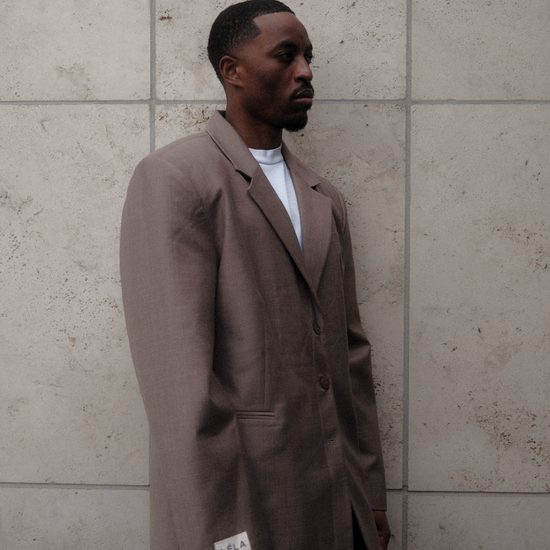 Side profile of male model wearing beige Carter Blazer with white LÉLA branded label visible on cuff, styled with dark denim against textured concrete wall