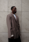 Side profile of male model wearing beige Carter Blazer with white LÉLA branded label visible on cuff, styled with dark denim against textured concrete wall