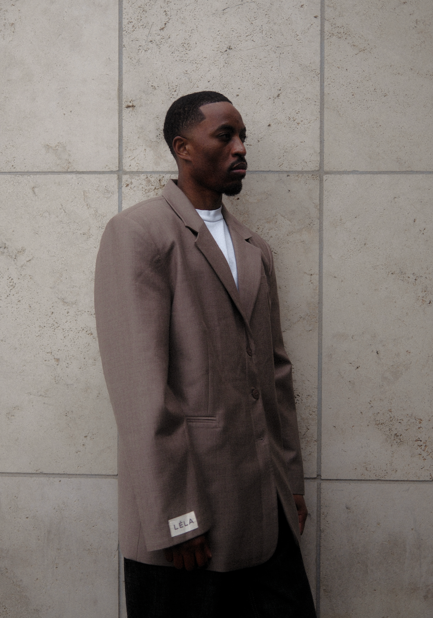 Side profile of male model wearing beige Carter Blazer with white LÉLA branded label visible on cuff, styled with dark denim against textured concrete wall