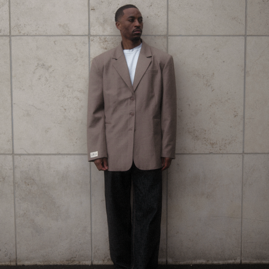 Male model wearing beige Carter Blazer with white t-shirt and dark washed denim against textured white concrete wall, showing oversized tailored fit