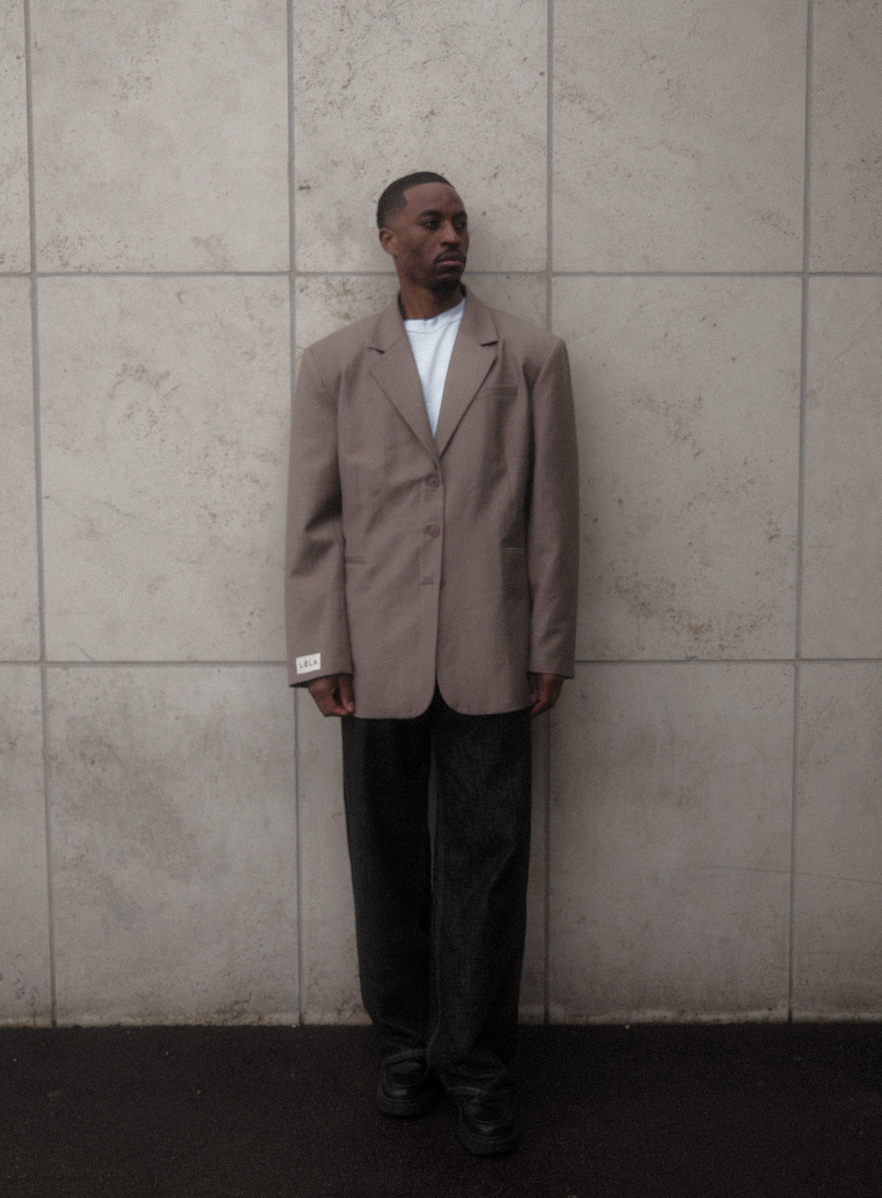 Male model wearing beige Carter Blazer with white t-shirt and dark washed denim against textured white concrete wall, showing oversized tailored fit
