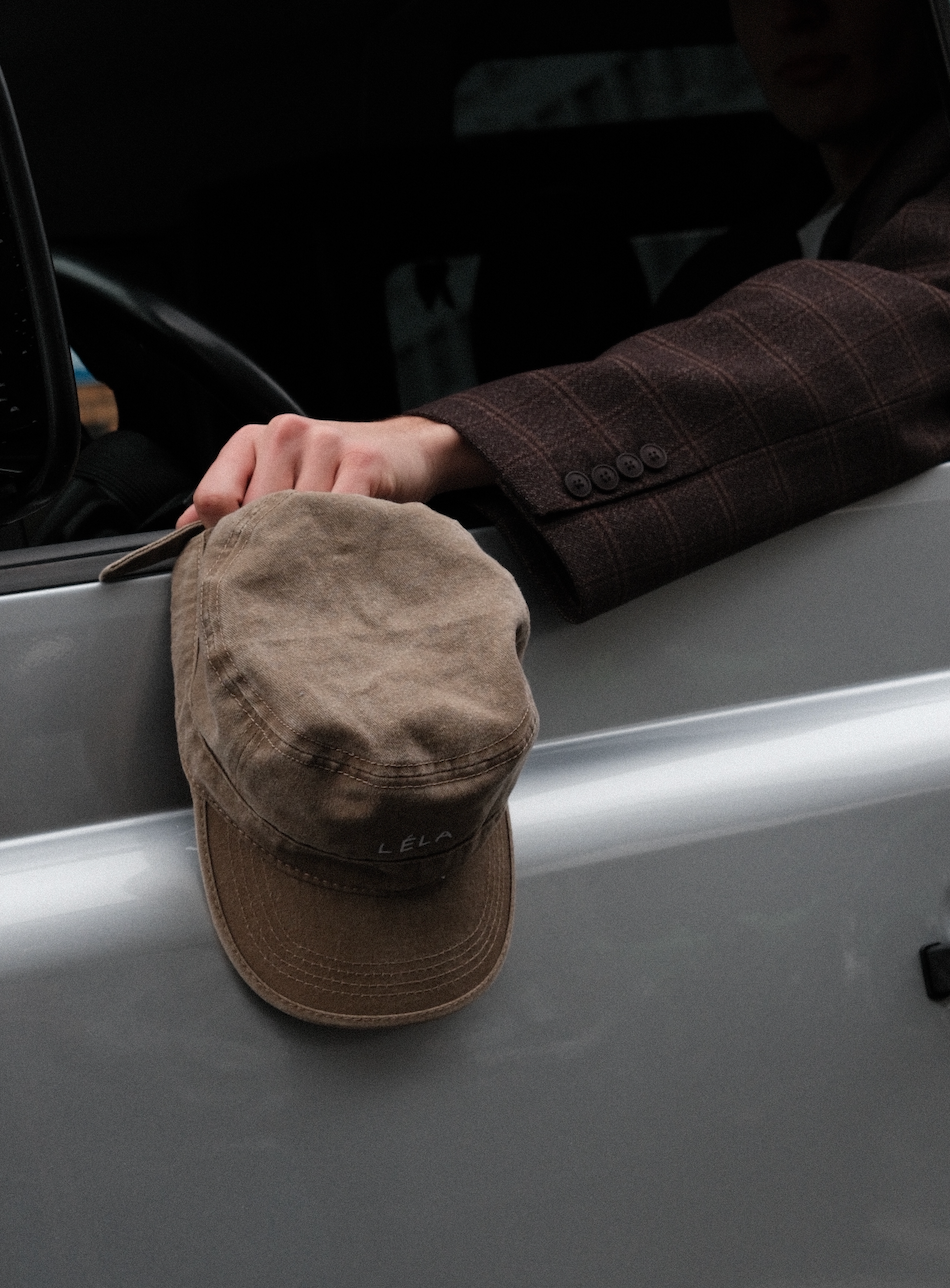 Top-down view of washed army green LÉLA cap showing embroidered logo detail and distressed cotton texture, held by hand in brown LÉLA Archer blazer